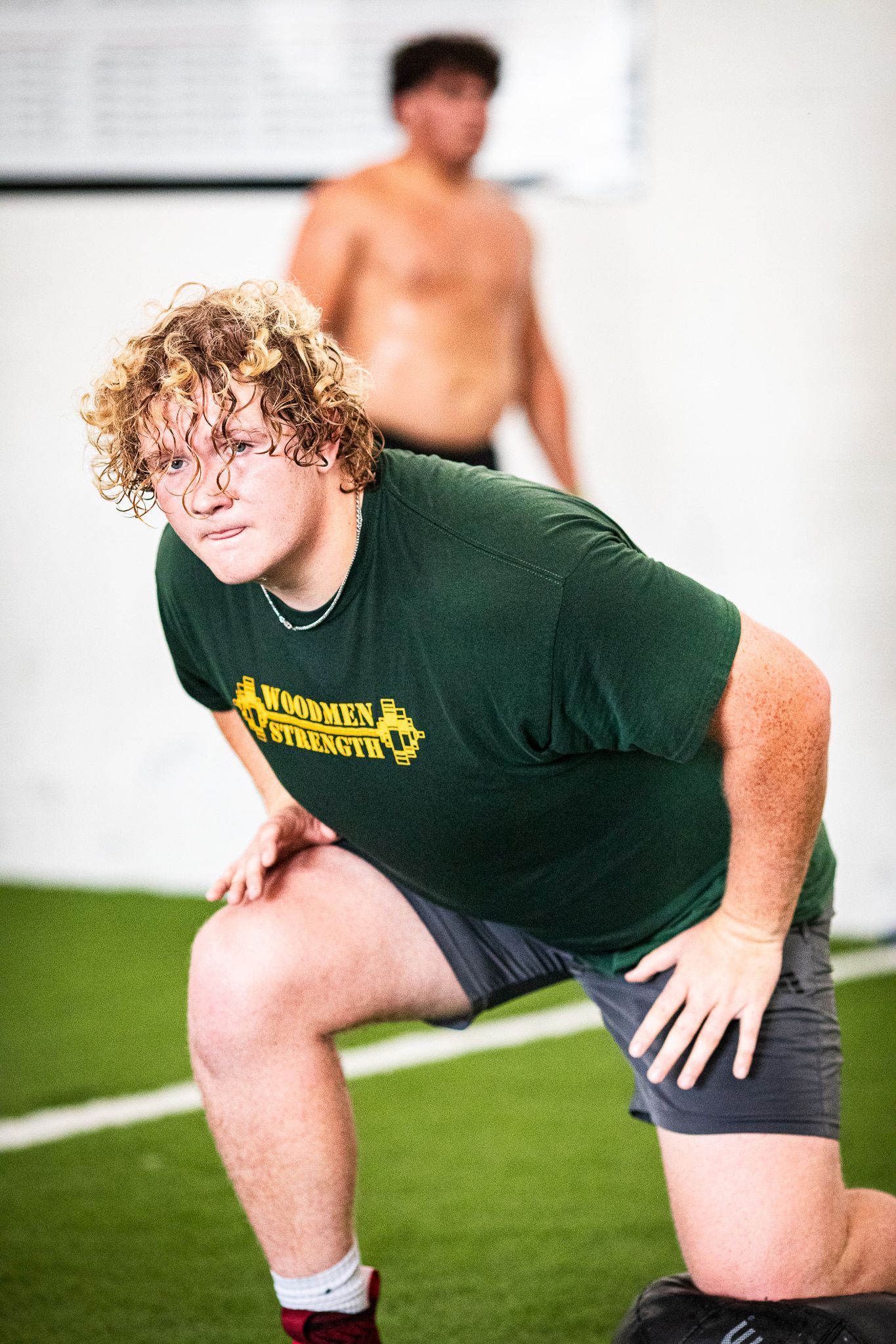 Young man in green shirt stretches on a tire. Another shirtless man in the background. Gym setting.