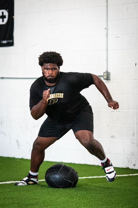 Athlete in black gear sprints toward a medicine ball on indoor turf.