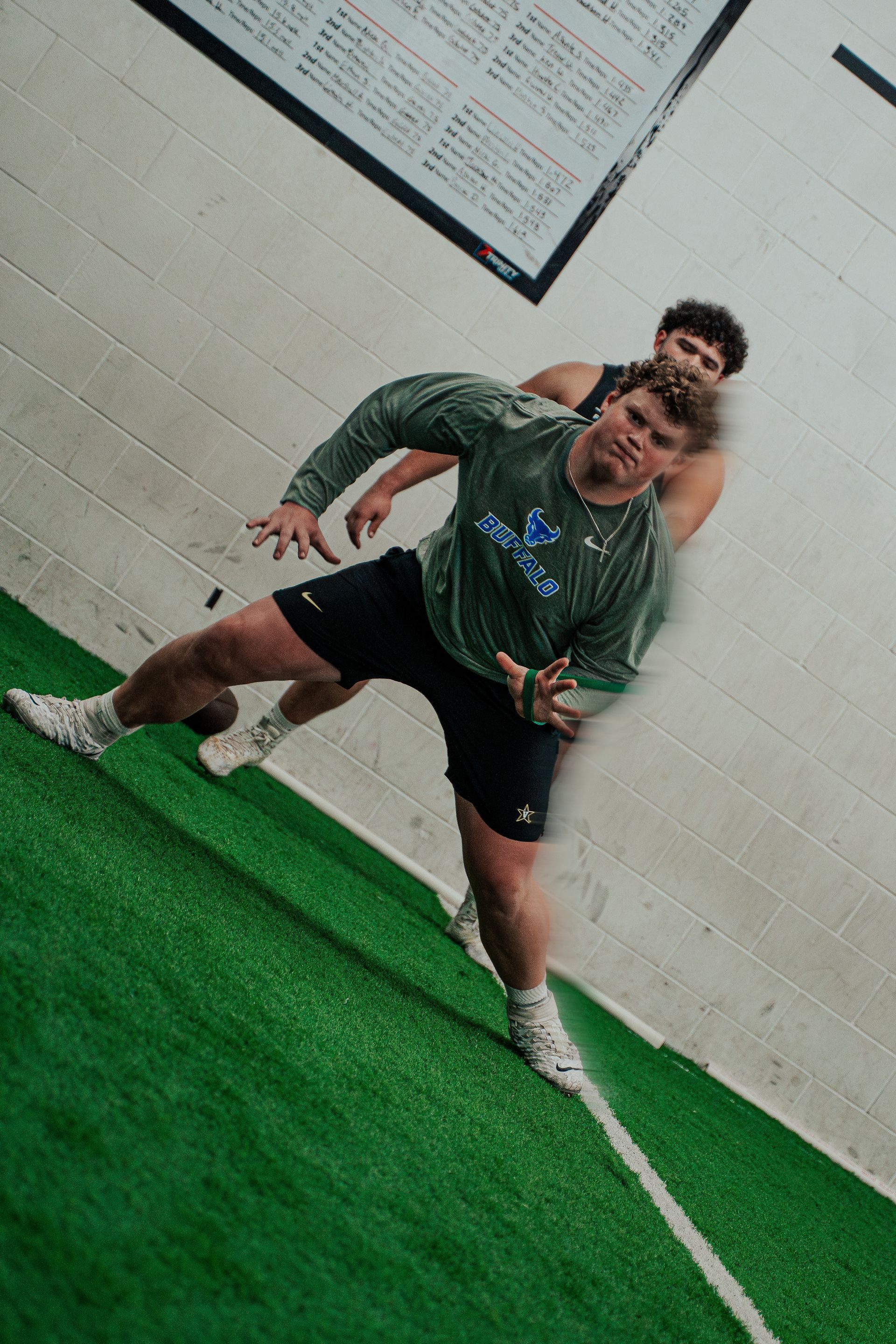 Two men exercising on a green turf track. One leans forward, arms out. The other stands behind him.