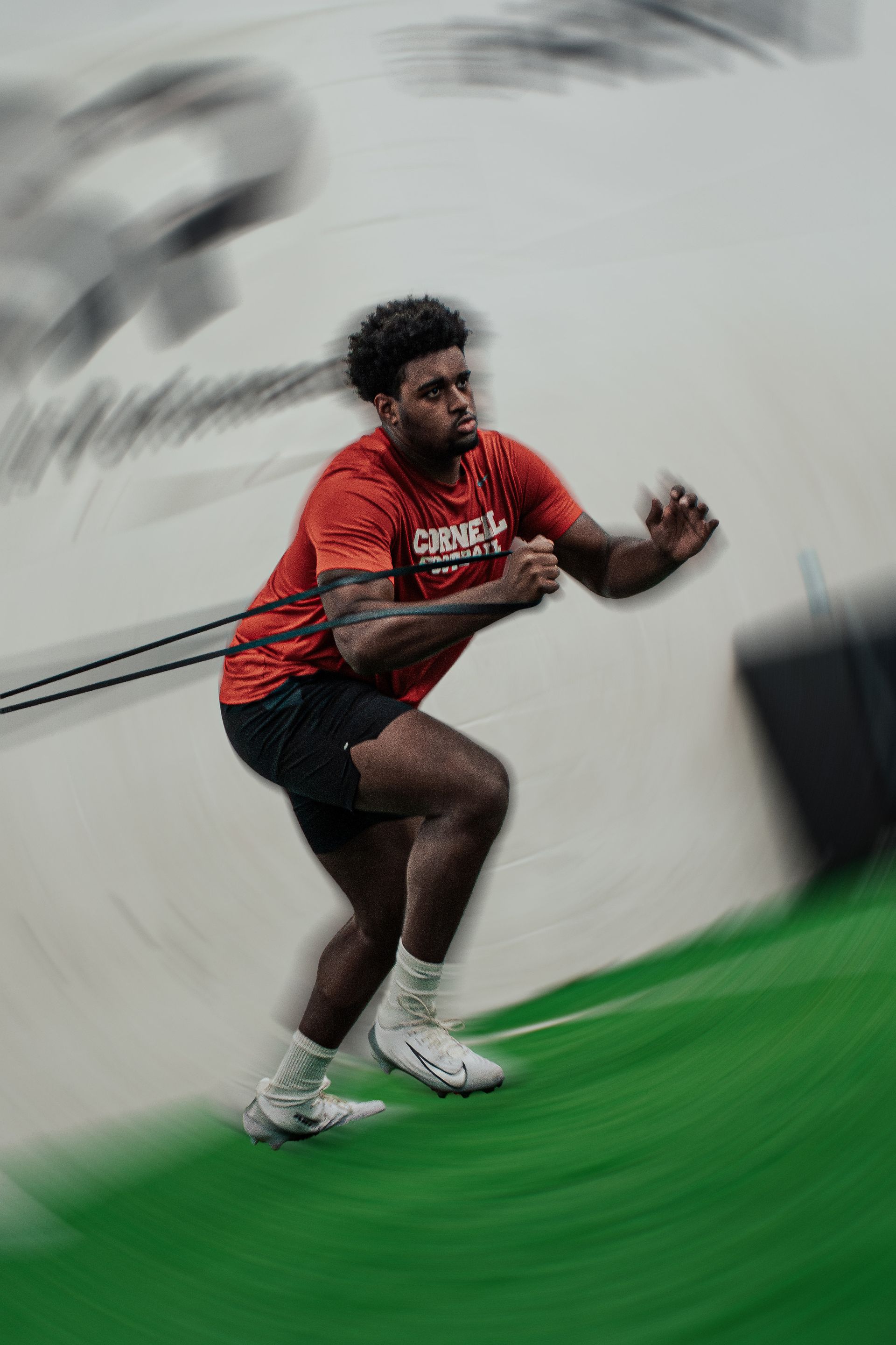 Athlete in red shirt sprints on green turf, pulling resistance band indoors.