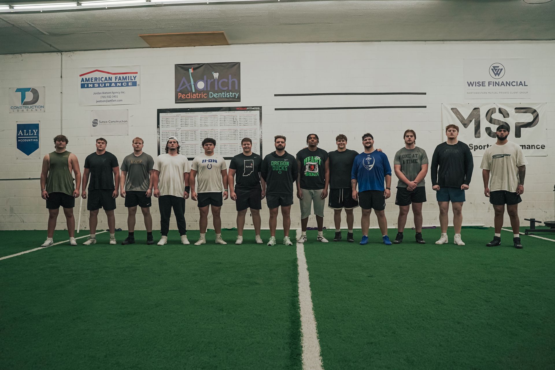 A group of men standing on green turf in a gym in front of a white wall.