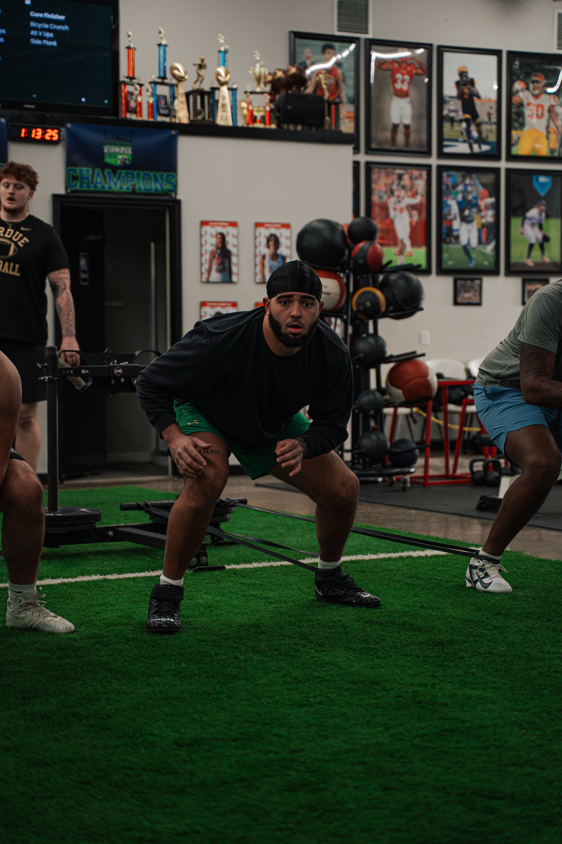 Three athletes in a gym doing an athletic drill. One person in focus has a black shirt and green shorts, performing a squat.