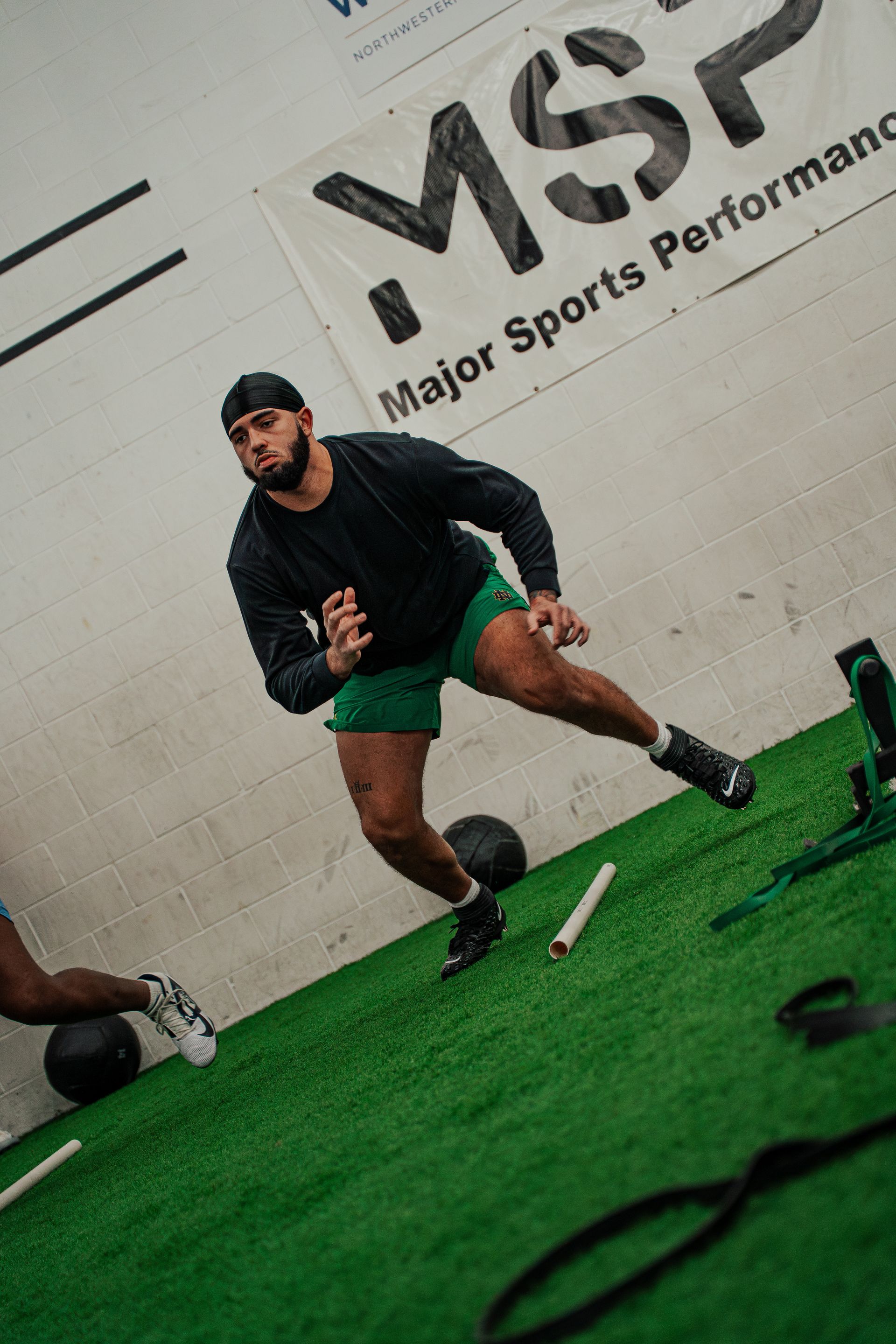 A man in athletic wear runs sideways on a turf field. Training facility with MSP banner in background.