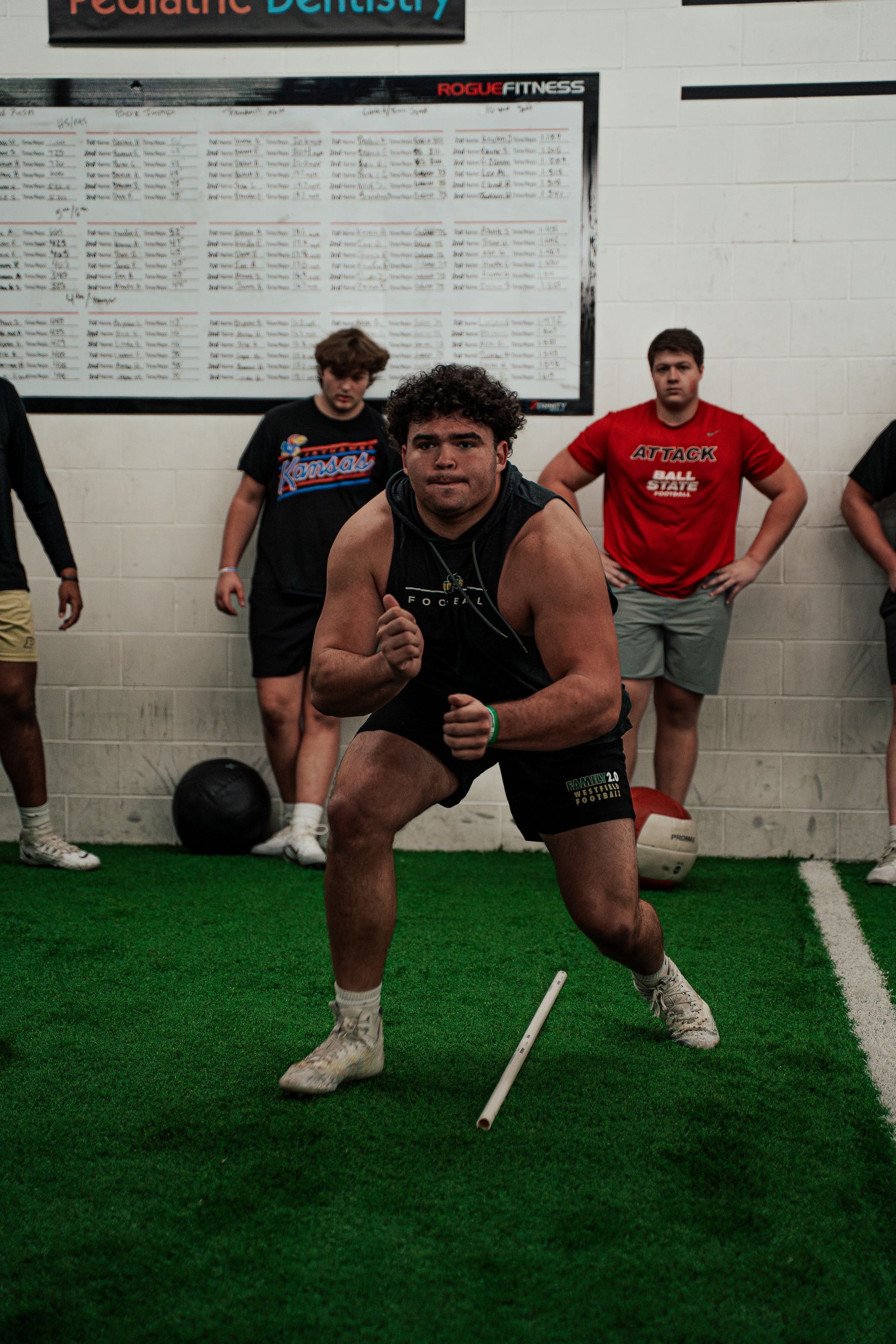 Athlete in workout attire, lunging with a marker on an indoor turf field; other people in the background.