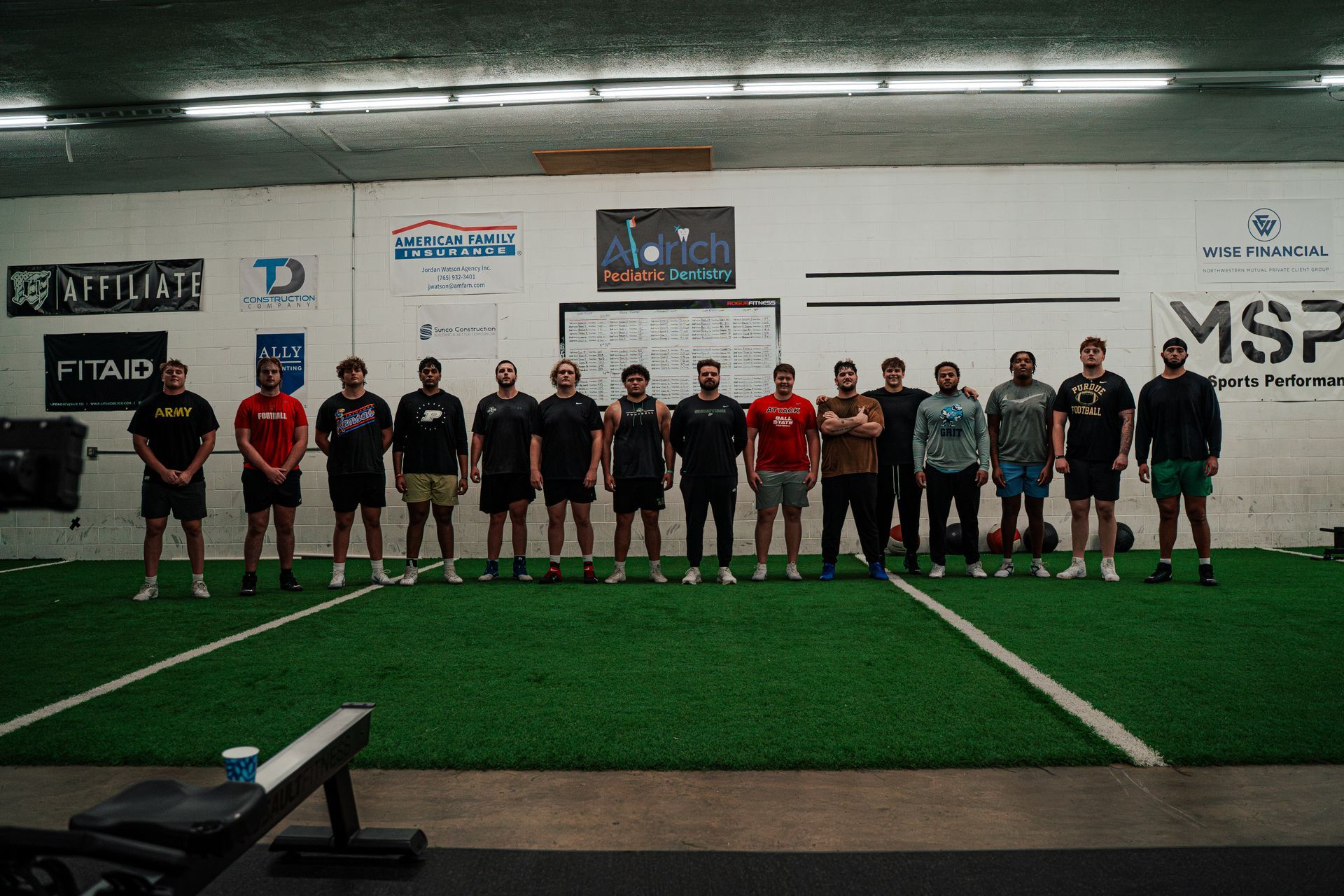 Group of men standing on a turf field in a gym. Several are wearing athletic clothes.