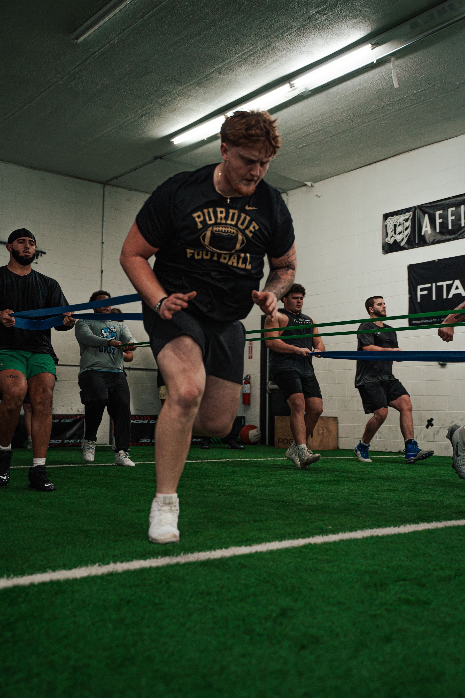Athlete in black Purdue shirt sprints forward, pulling against a resistance band in a gym.