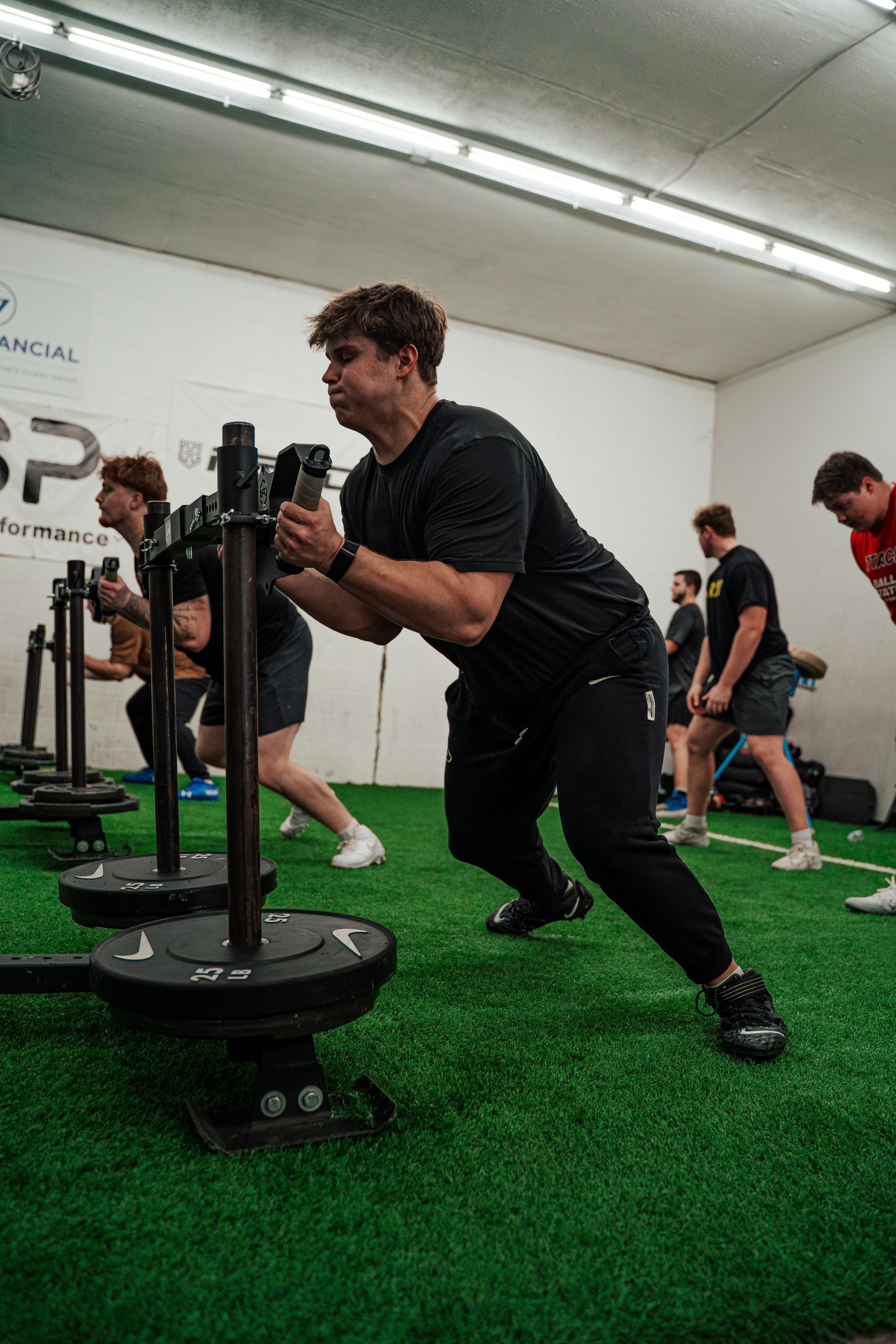 People pushing weighted sleds on a turf floor, in a gym. Black outfits, focused expressions.