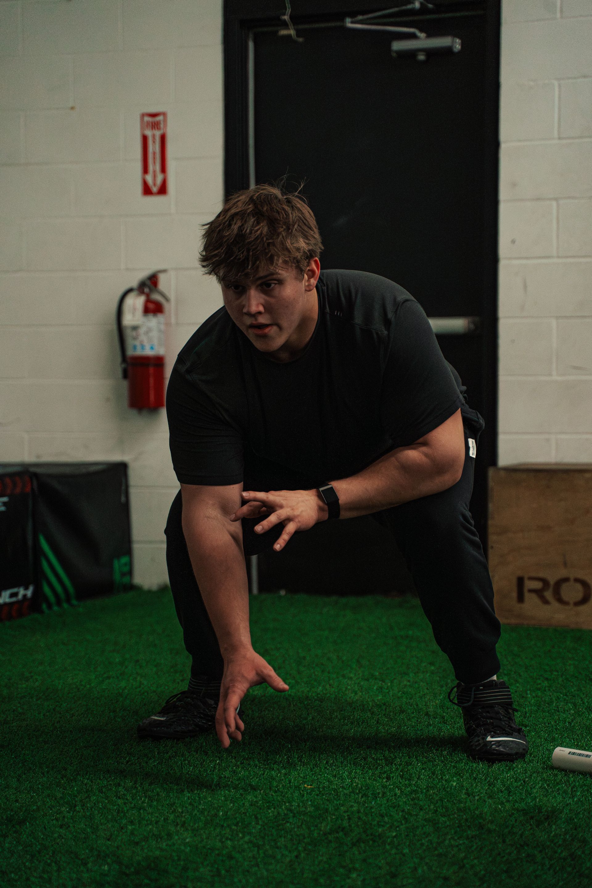 Man in black workout gear crouches, hands forward, on green turf, gym setting.