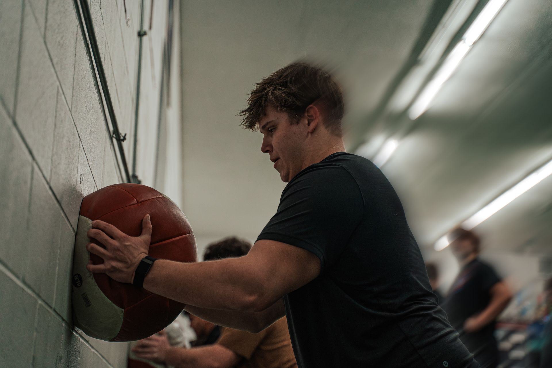Man doing wall ball exercise in a gym. He is wearing a black shirt and a watch.