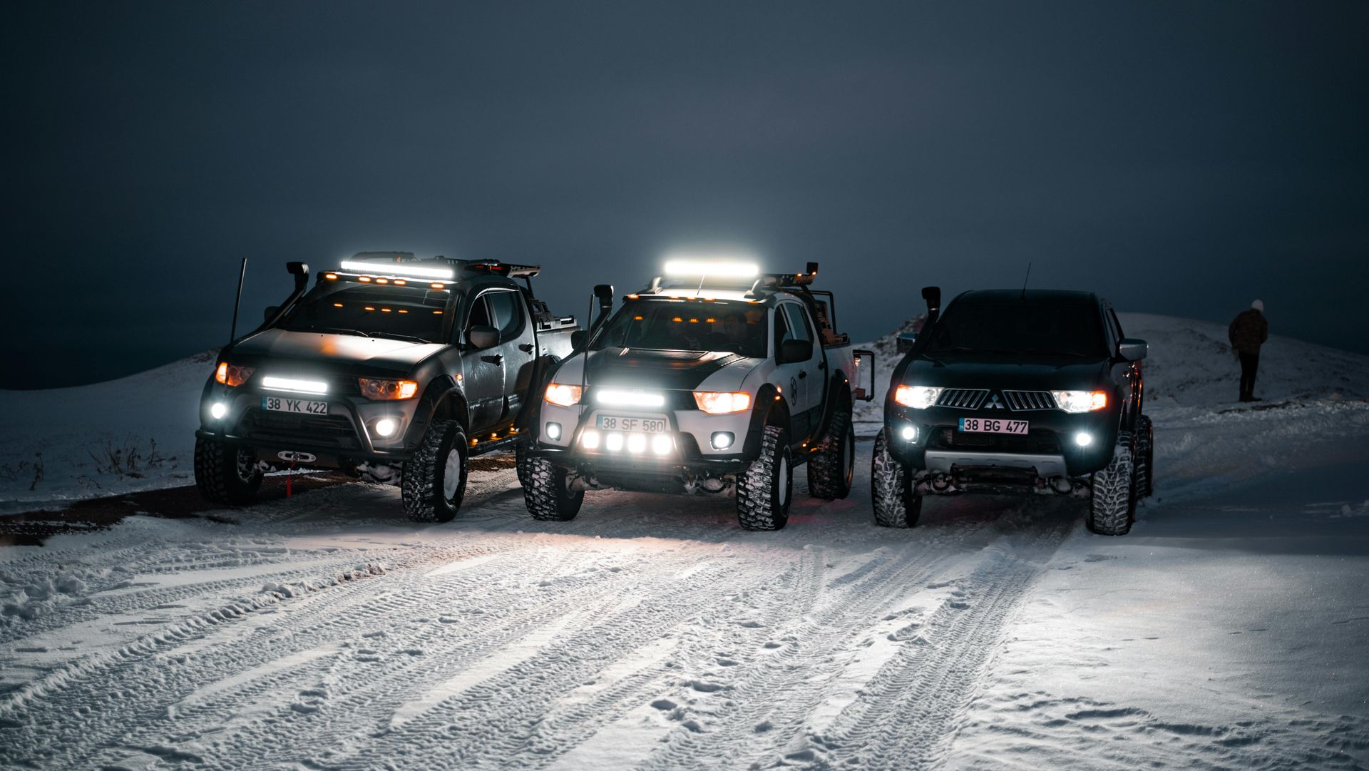 Three modified off-road vehicles with bright lights on a snowy road at night. One person stands nearby.