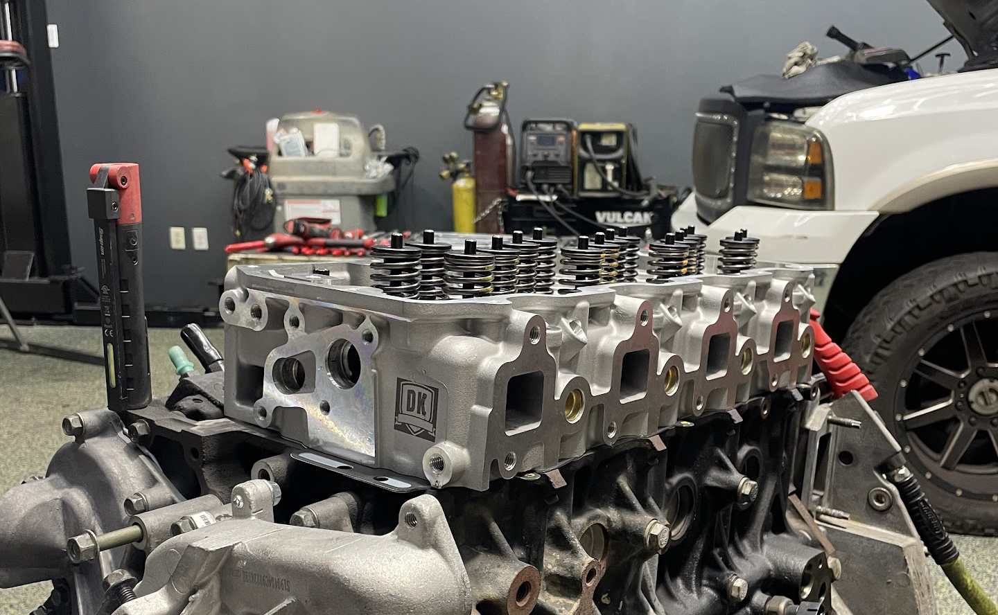 Engine block with a new cylinder head in a garage setting, next to a white truck.