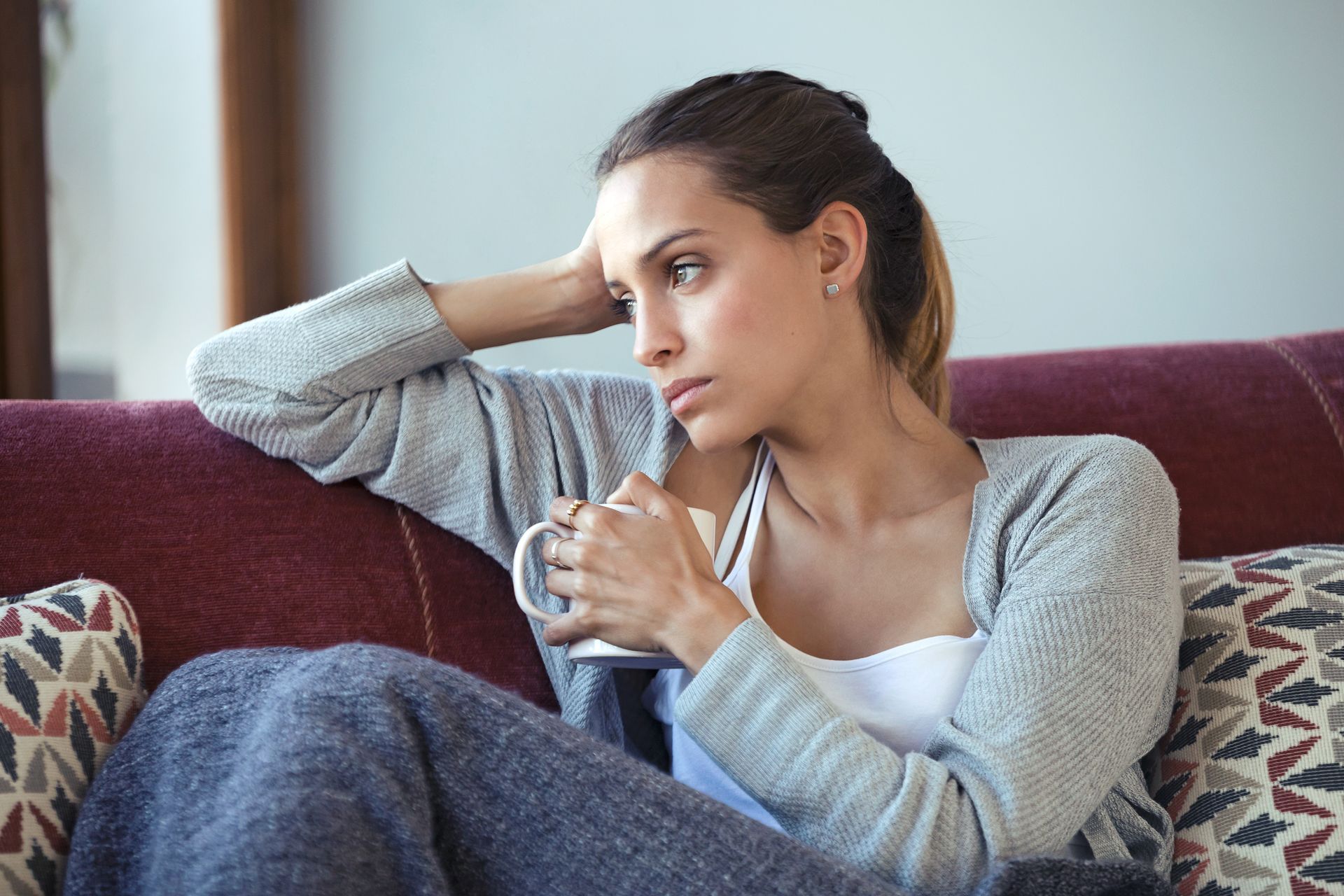 Woman sitting on a couch, holding a mug and looking away thoughtfully in a cozy room