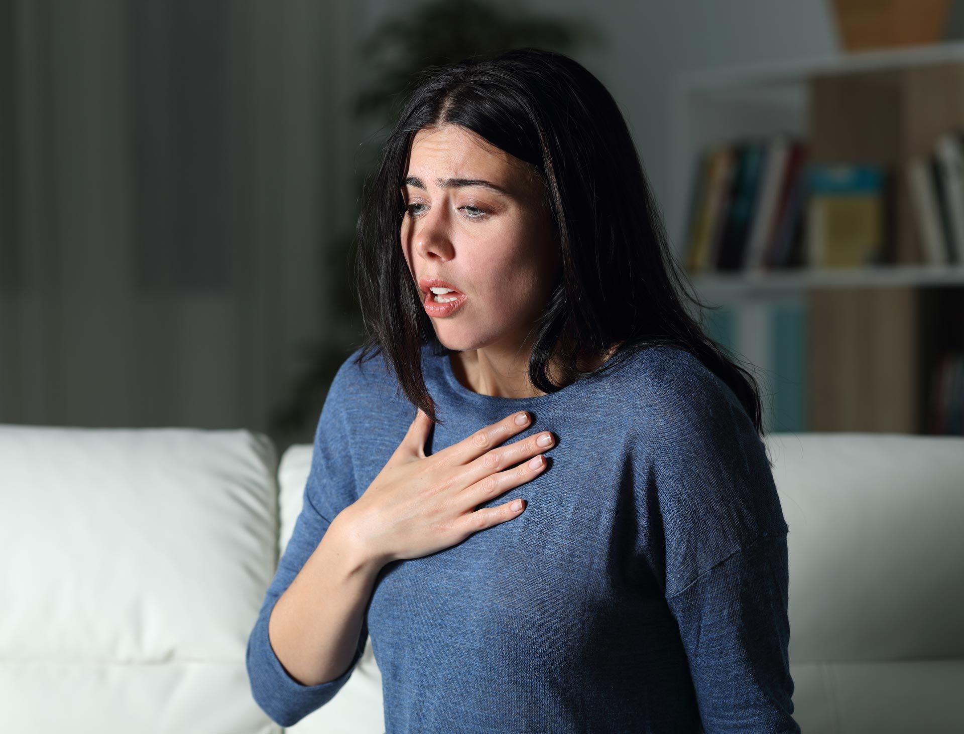 Woman in a blue sweater clutching her chest and looking distressed on a couch.