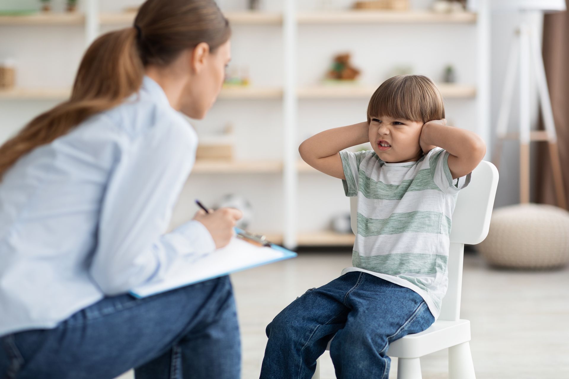 Counselor talking with a child in a bright therapy room while the child sits with hands over ears