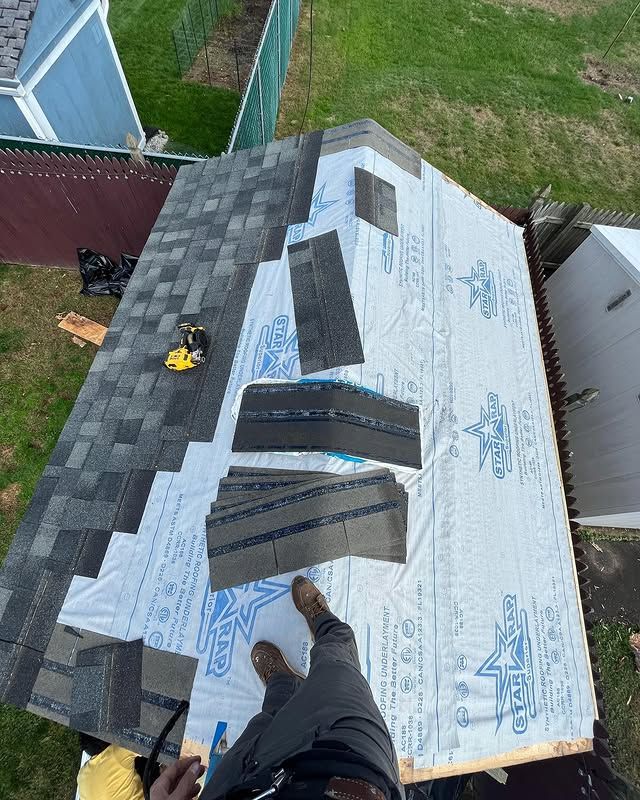 A man is standing on top of a roof with shingles on it.