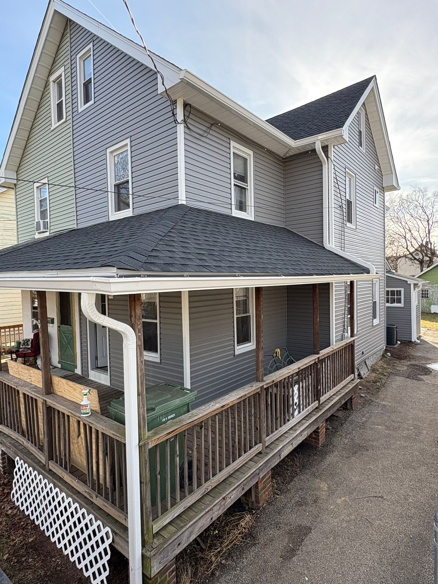 Two-story house with gray siding, a porch, and black roof under a blue sky.