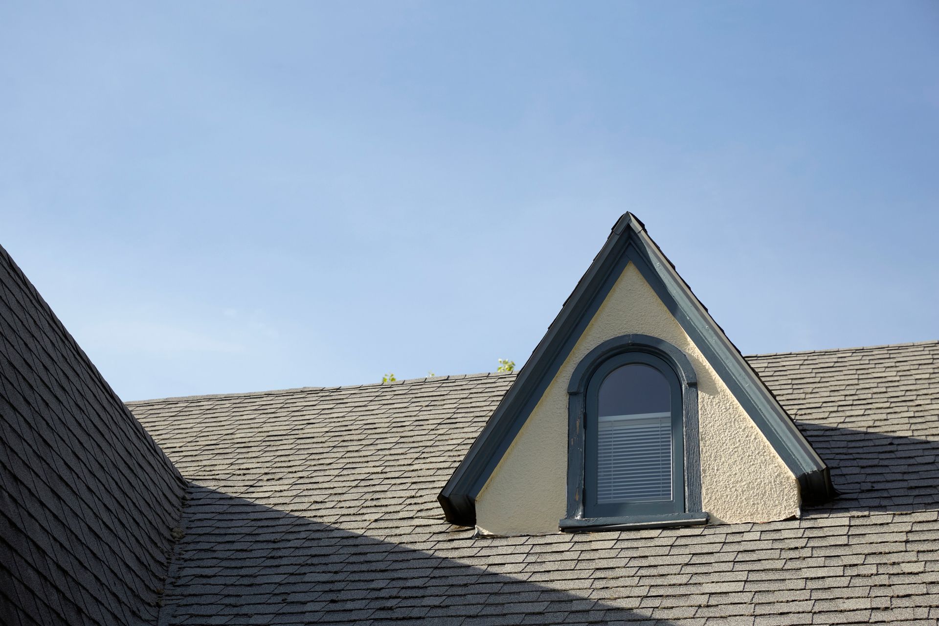 Roof with gray shingles and a small triangular dormer window against a clear blue sky. Roof with gray shingles and a small triangular dormer window against a clear blue sky.
