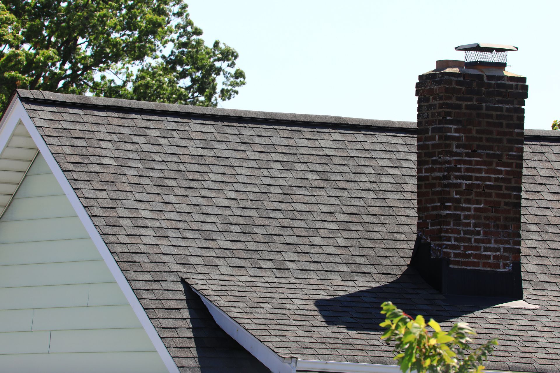 Close-up of a house roof with dark shingles and a brick chimney under clear sky.