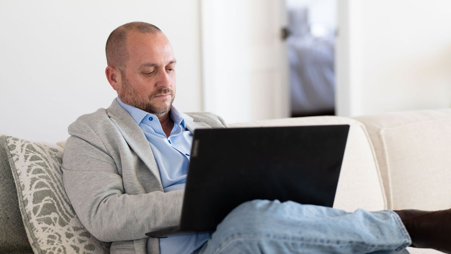 Man using a laptop while sitting on a couch. He wears a light grey jacket, light blue shirt, and jeans.