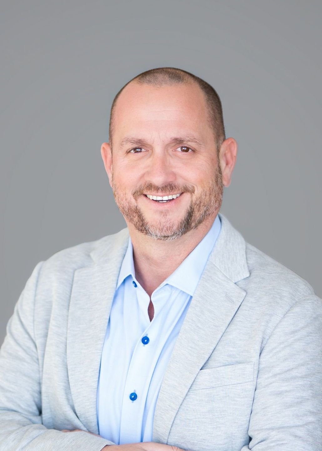 Man with a beard smiling, wearing a light blue shirt and gray blazer against a gray backdrop.