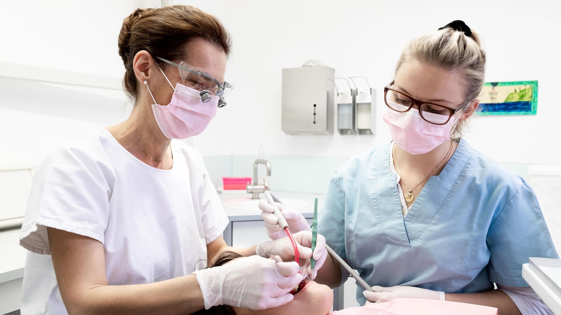 Zwei Zahnärzte in OP-Kleidung, Masken und Schutzbrillen behandeln einen Patienten in einer Praxis.