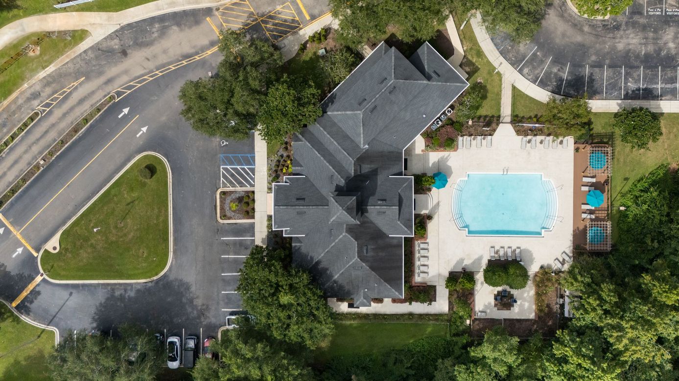 Aerial view of a clubhouse complex with a dark roof, surrounding parking, a blue swimming pool, and lush green landscaping.