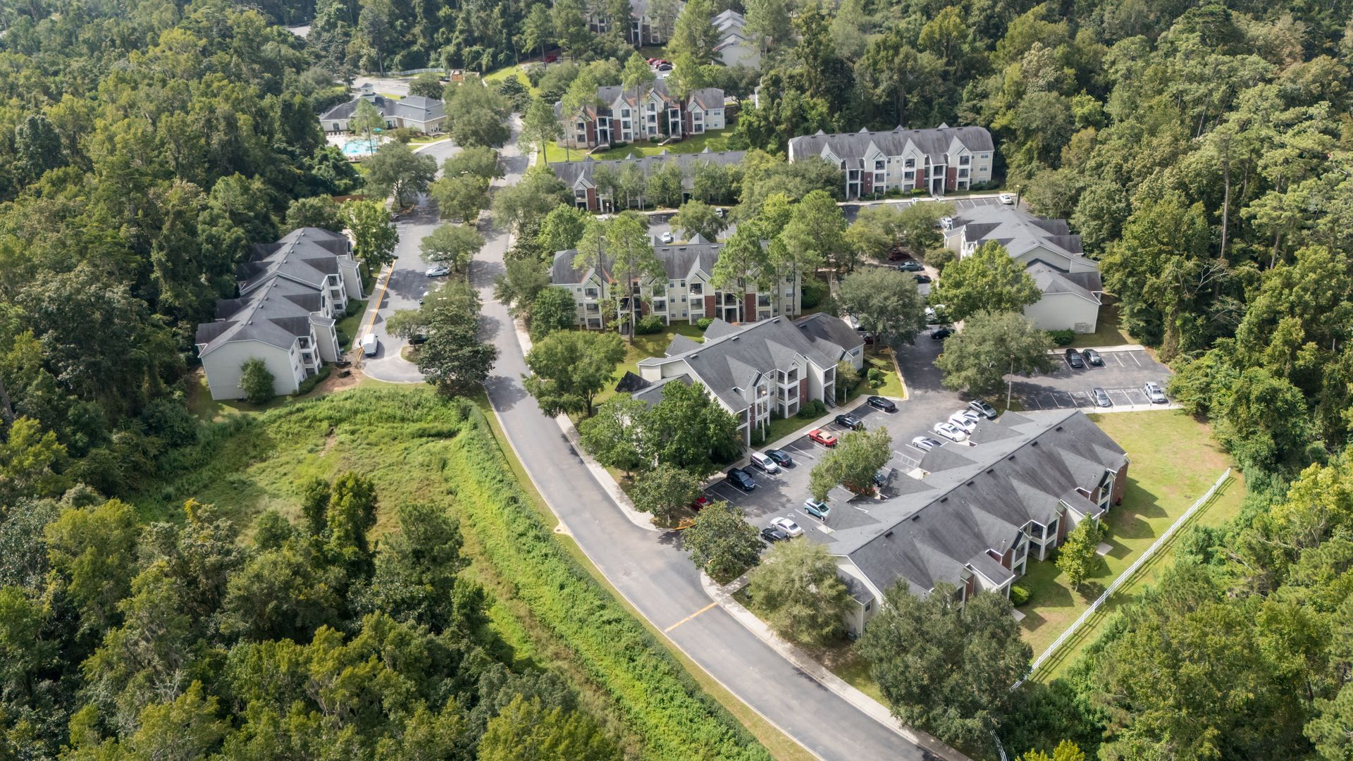 Aerial view of a residential complex surrounded by lush green trees, with buildings and a winding road visible.