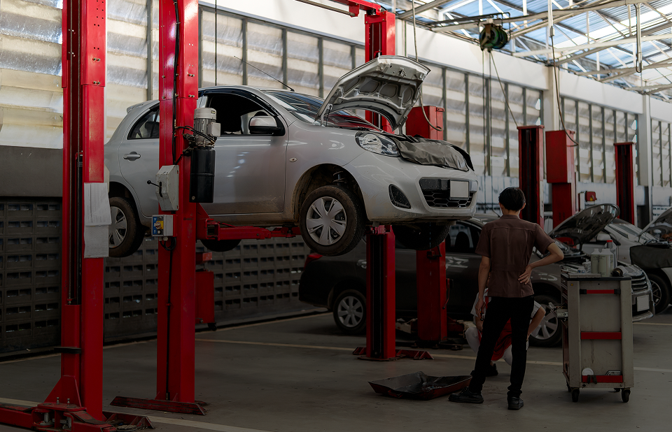 Car on a lift in a garage, mechanic standing nearby, looking at the vehicle with open hood. | Lambert's Auto Repair