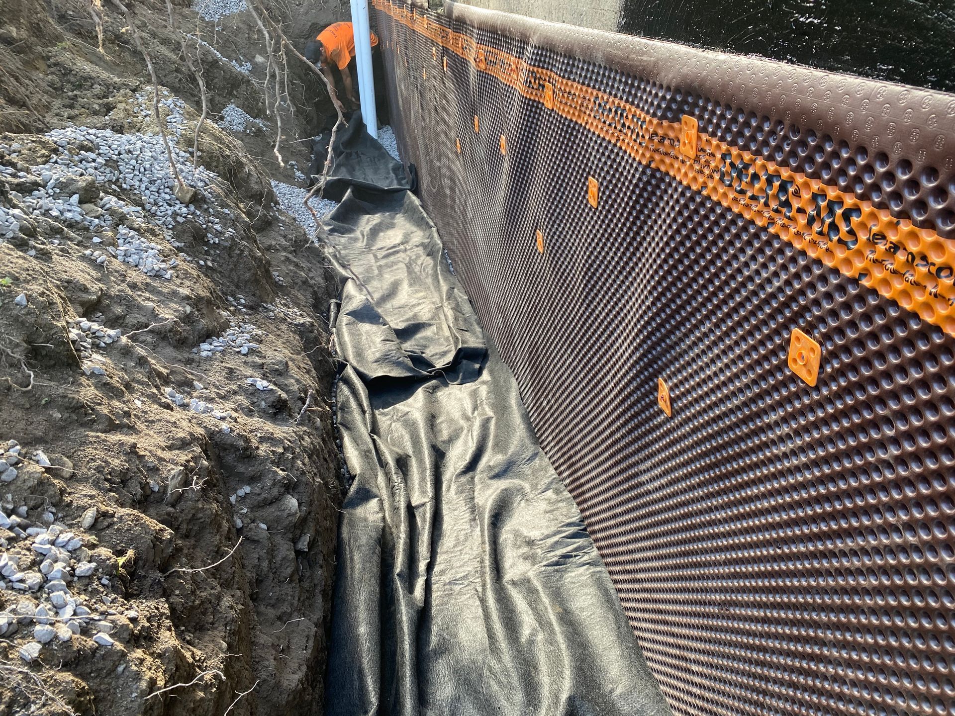 Installation du système de drainage : tranchée avec géotextile noir, membrane plastique et tuyau blanc.