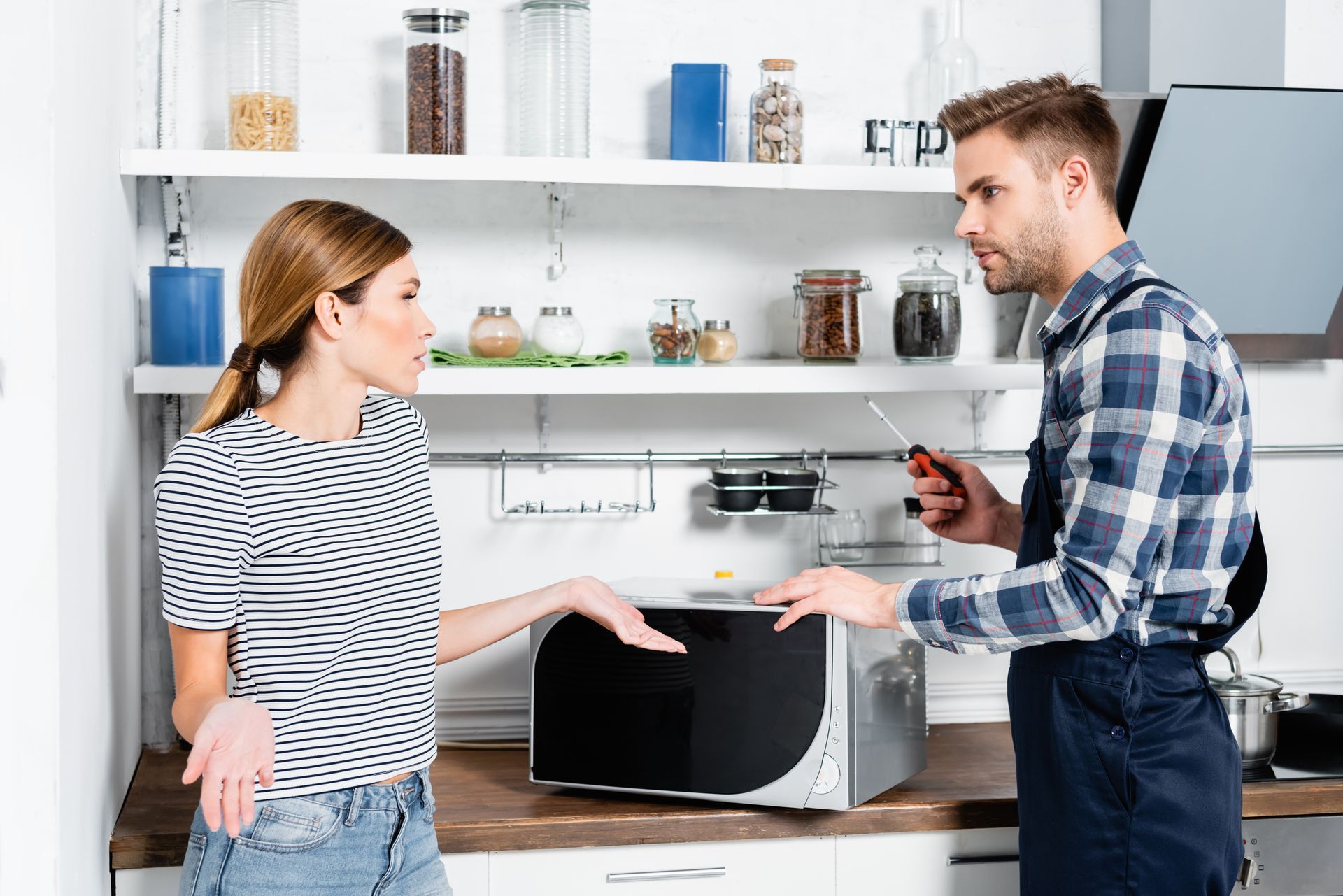 A man is fixing a microwave in a kitchen while a woman looks on.