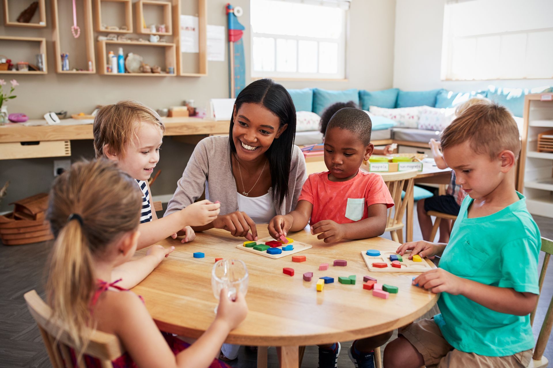 A teacher and four children sit at a round wooden table, working together on colorful shape-sorting puzzles in a classroom.