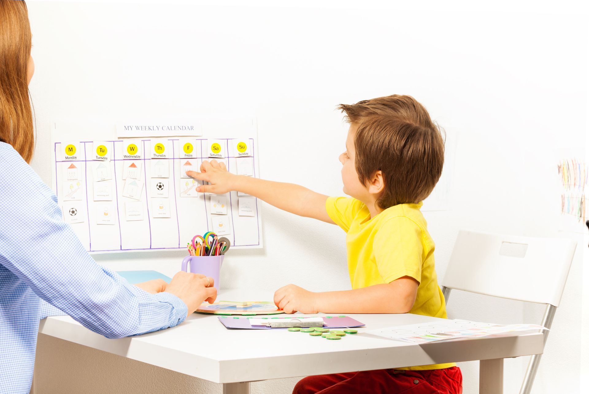 A child pointing at a weekly calendar on the wall while sitting at a table with an adult.