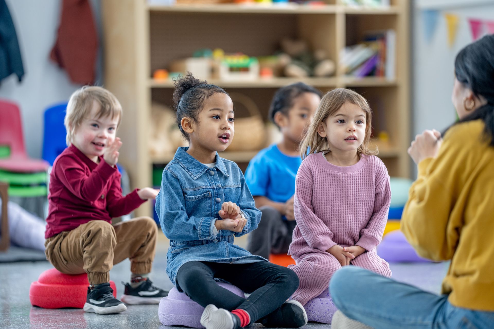 A small group of preschool children sit on the floor in front of their teacher.