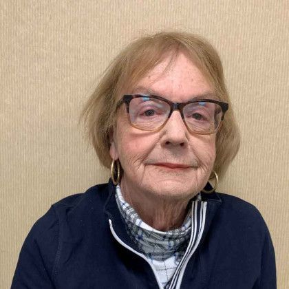 A woman wearing glasses and a blue jacket is sitting in front of a wall.