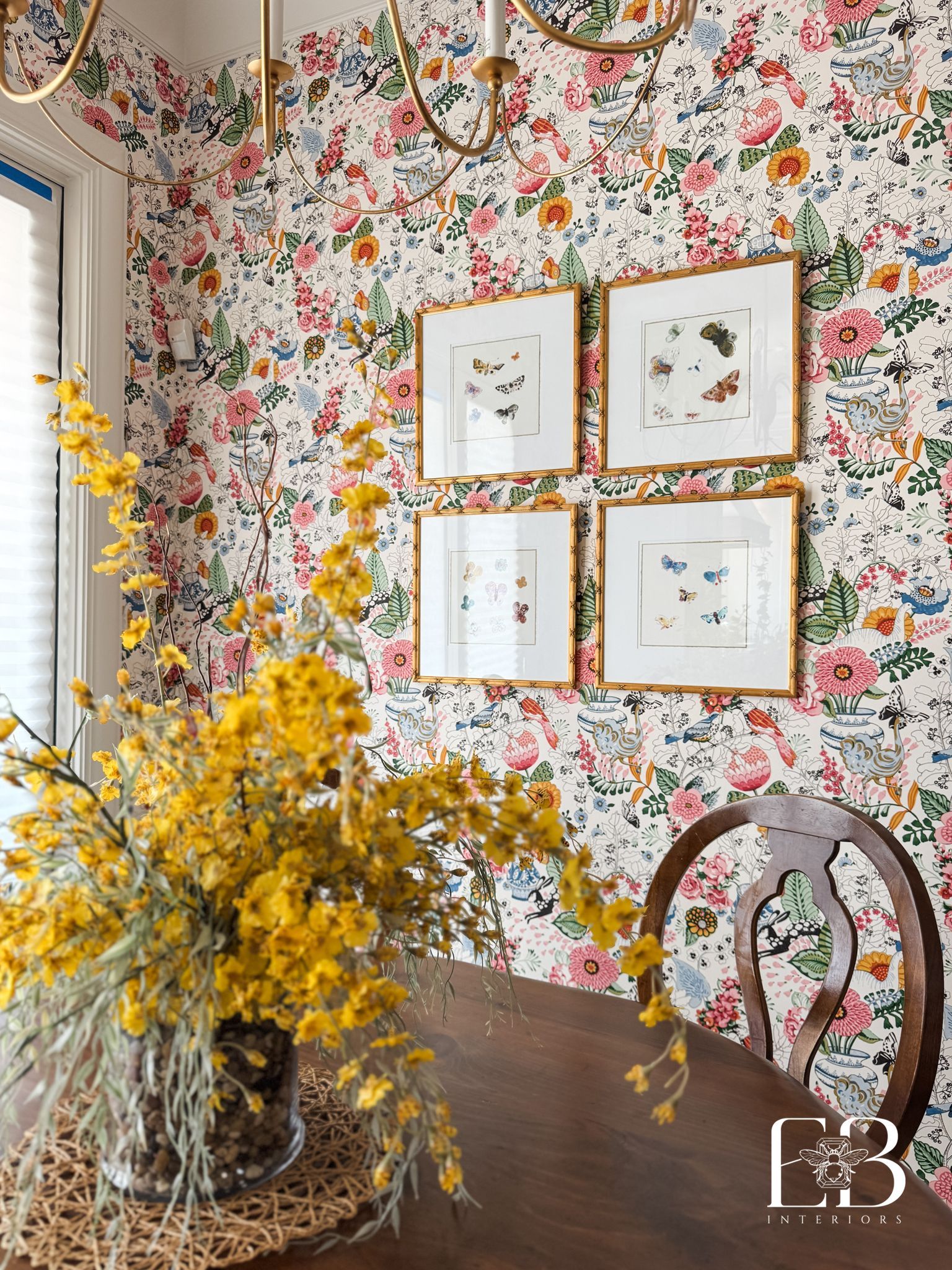 Dining room with floral wallpaper, framed art, yellow flowers on a table, and a wooden chair.