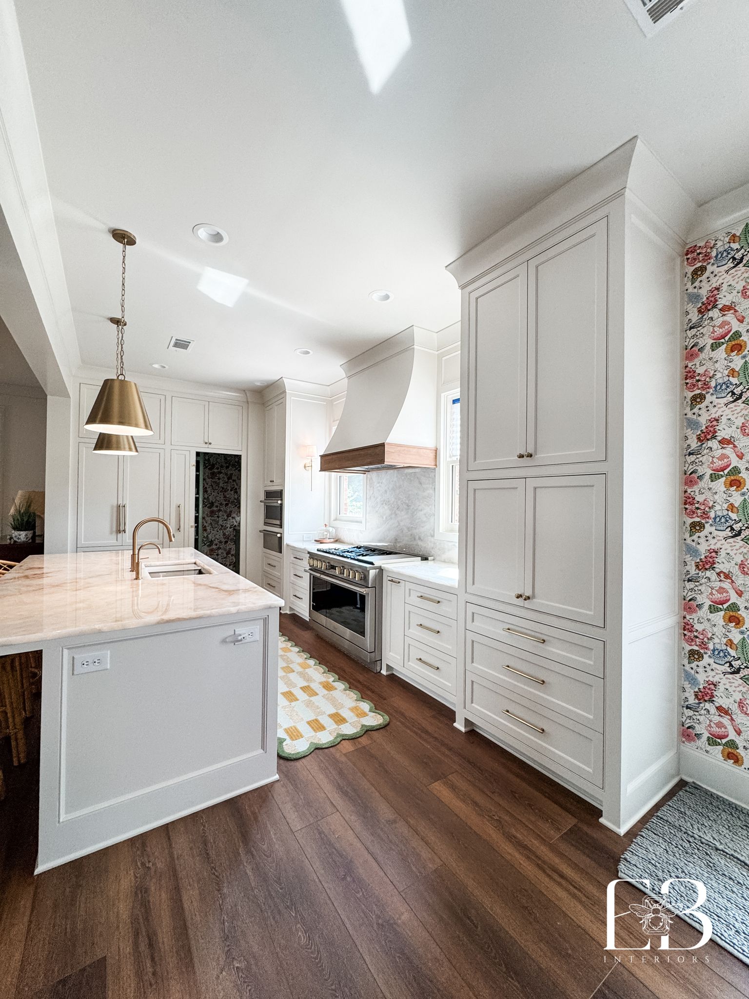 Bright white kitchen with a wooden floor, island, and cabinets, accented with gold fixtures and floral wallpaper.