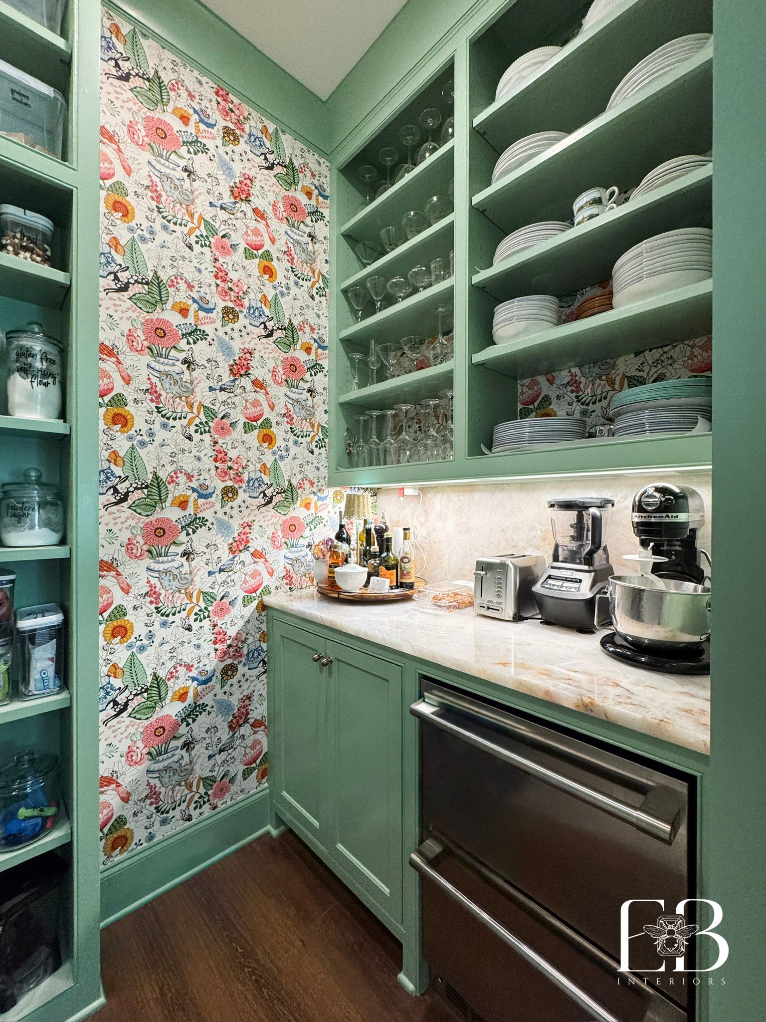 A mint-green pantry with floral wallpaper, shelves holding dishes, and a stainless steel appliance.