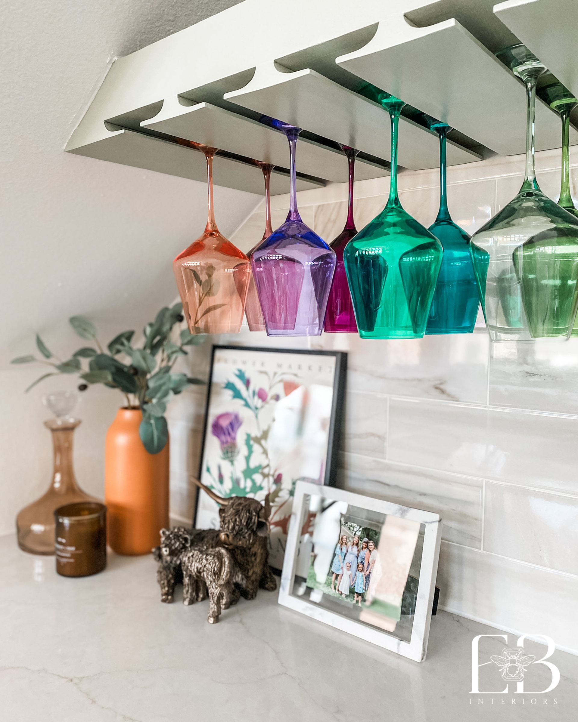 Wine glasses in rainbow colors hang under a white rack. Kitchen counter with decor.