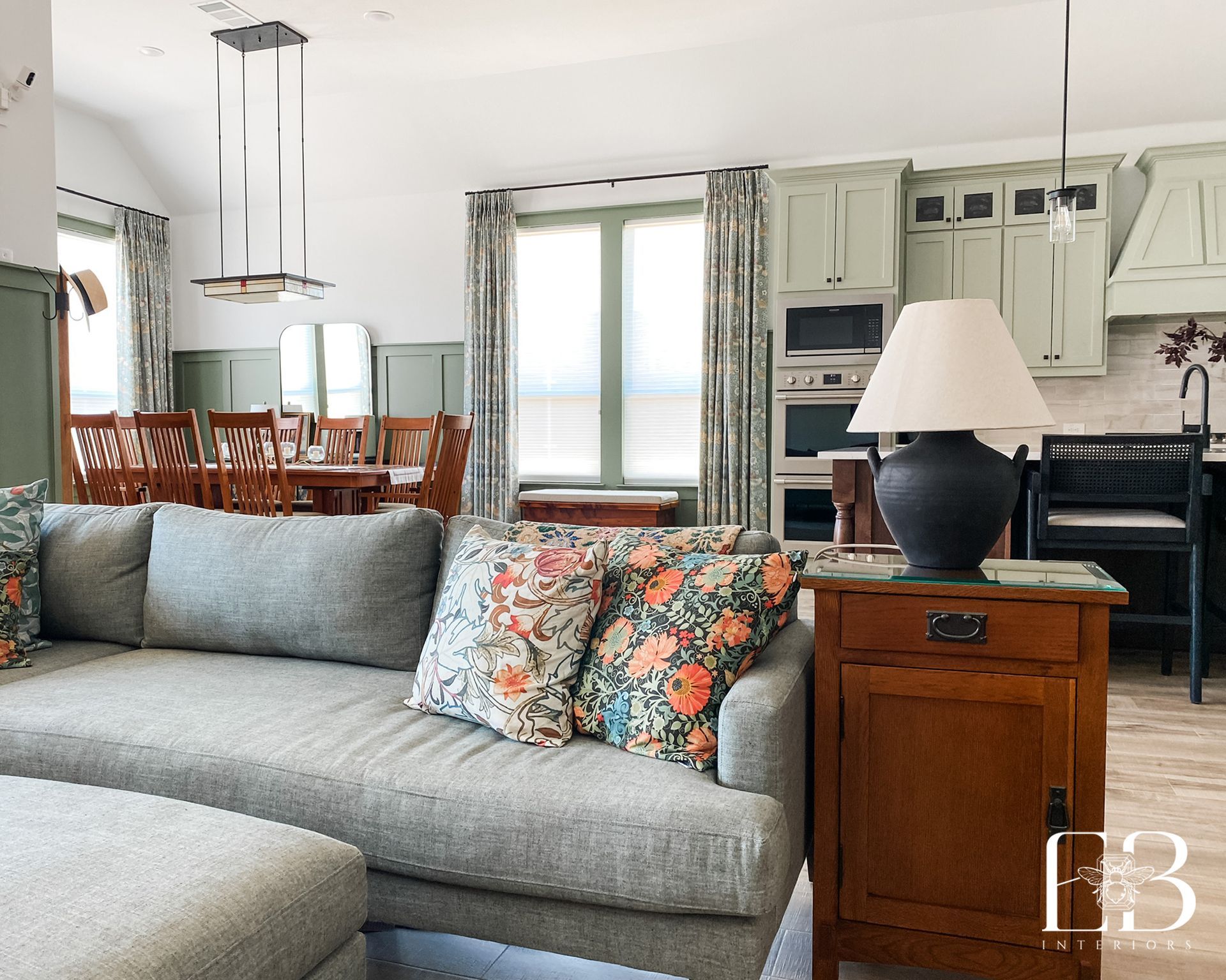 Living room with a gray sofa, floral pillows, wooden furniture, and green kitchen cabinets.