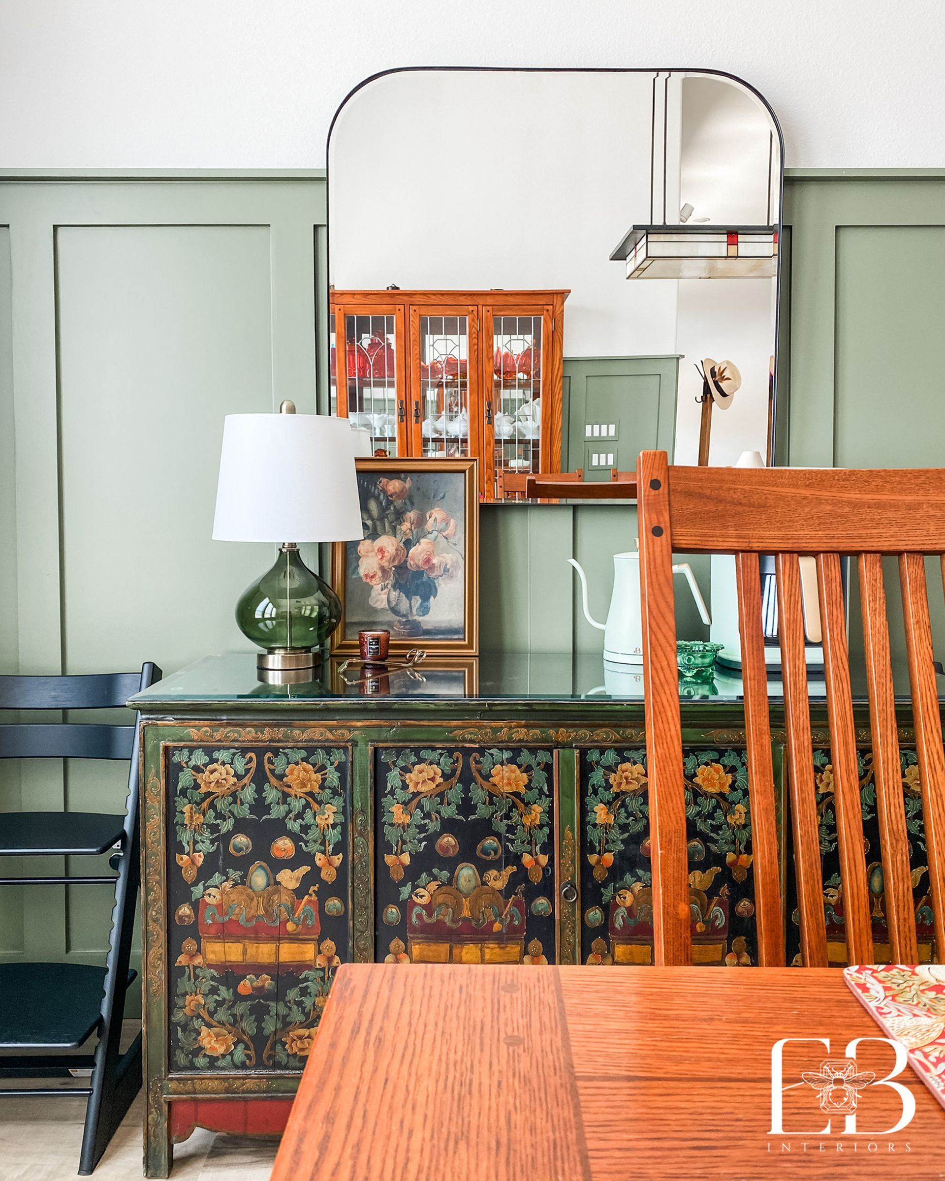 Dining area with green wainscoting, vintage floral credenza, and arched mirror styled by Emerald Bee Interiors.