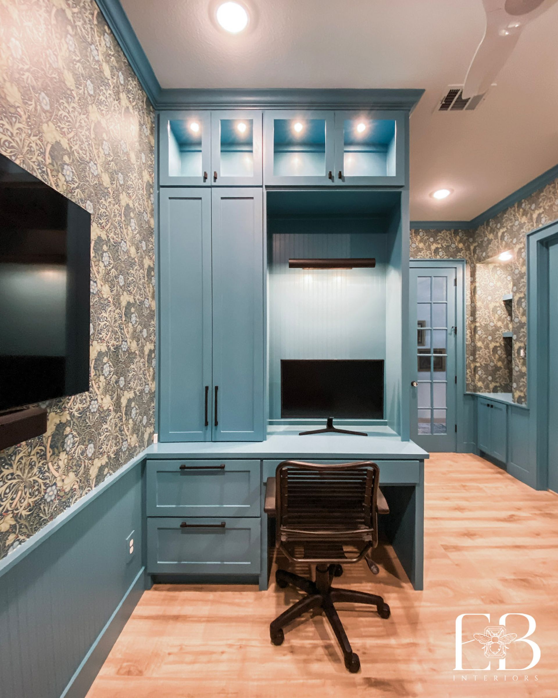Blue home office with built-in desk, cabinets, TV, floral wallpaper, and a black office chair.