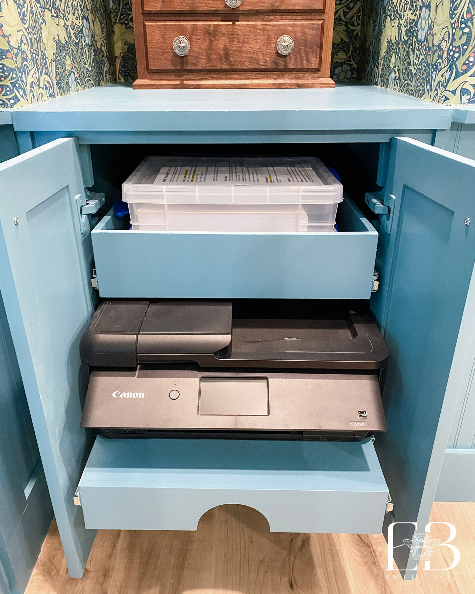 Blue cabinet with open doors, holding a clear storage bin, two printers. Wooden chest on top.