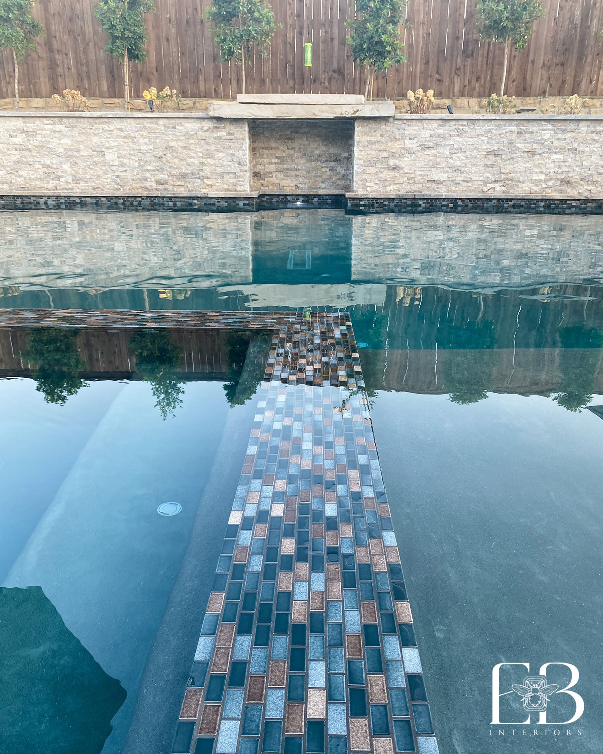 Pool with stone walls, decorative tile path, trees, and wooden fence.