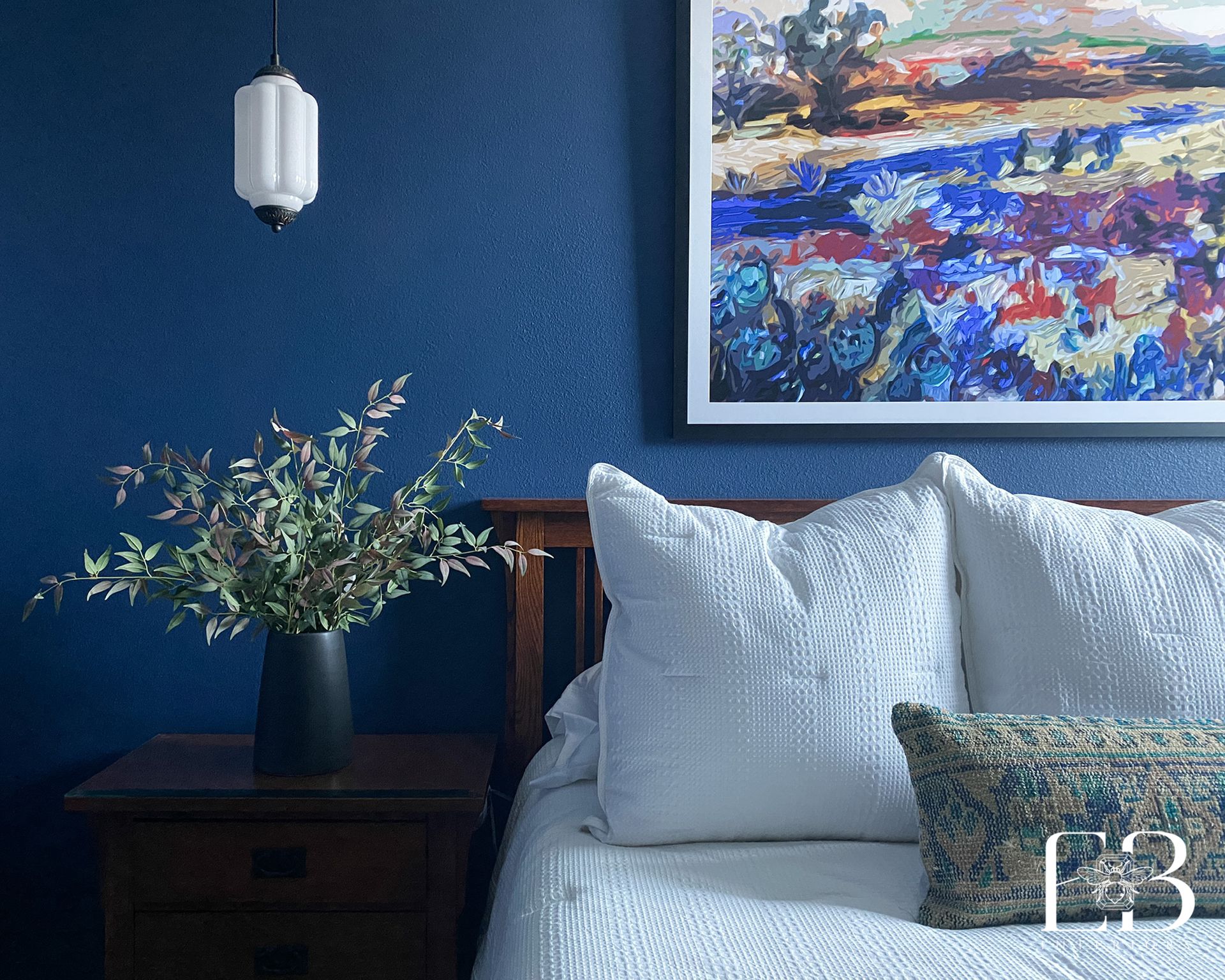 Bedroom with blue textured wall, art, white bedding, nightstand, and vase of greenery.