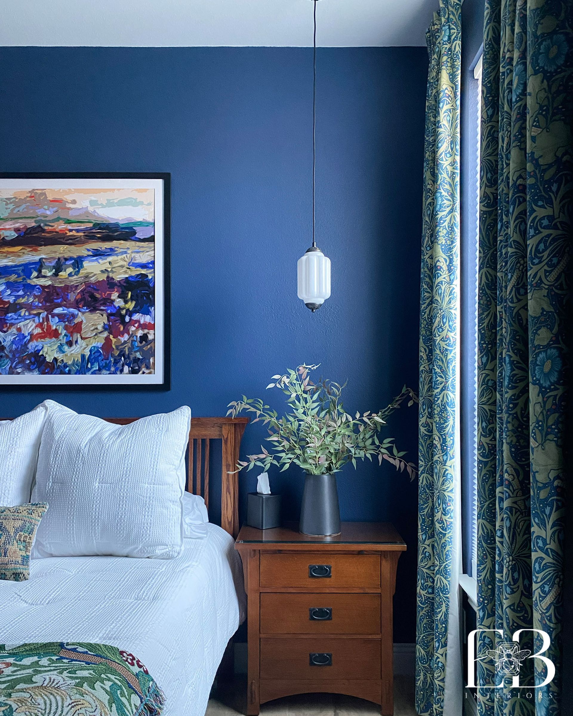 Bedroom with blue walls, art, and wooden nightstand; bed with white bedding, patterned curtains.