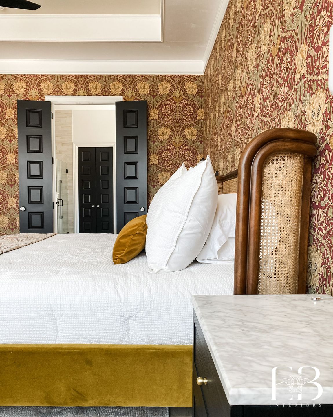 Primary bedroom with cane headboard, white bedding, and rich patterned wallpaper designed by Emerald Bee Interiors.