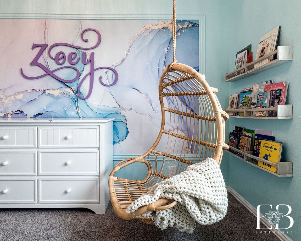 Girls’ bedroom with watercolor mural wall, rattan hanging chair, and lavender name sign by Emerald Bee Interiors.