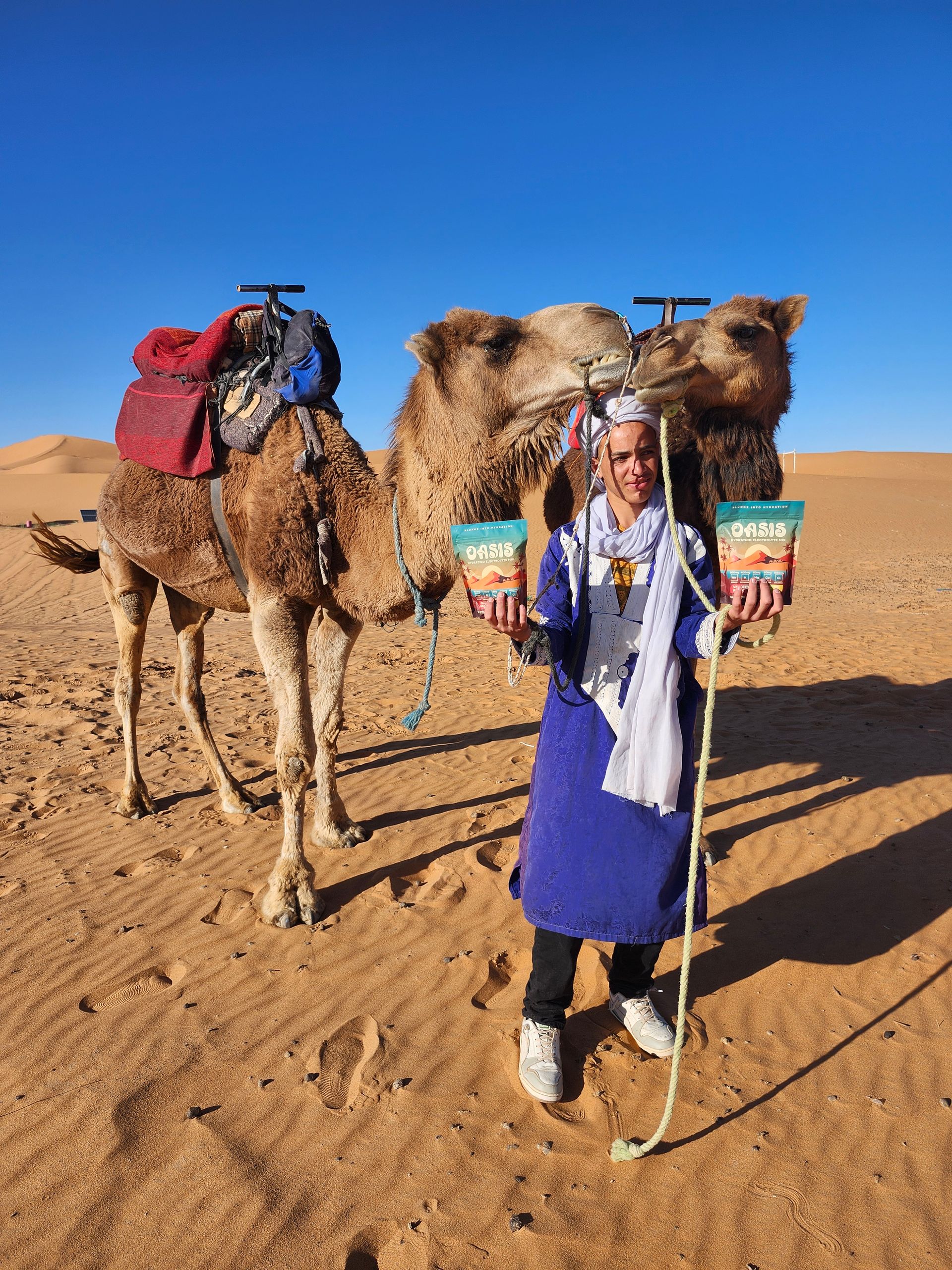 A woman is standing in front of two camels in the desert holding buckets of popcorn.