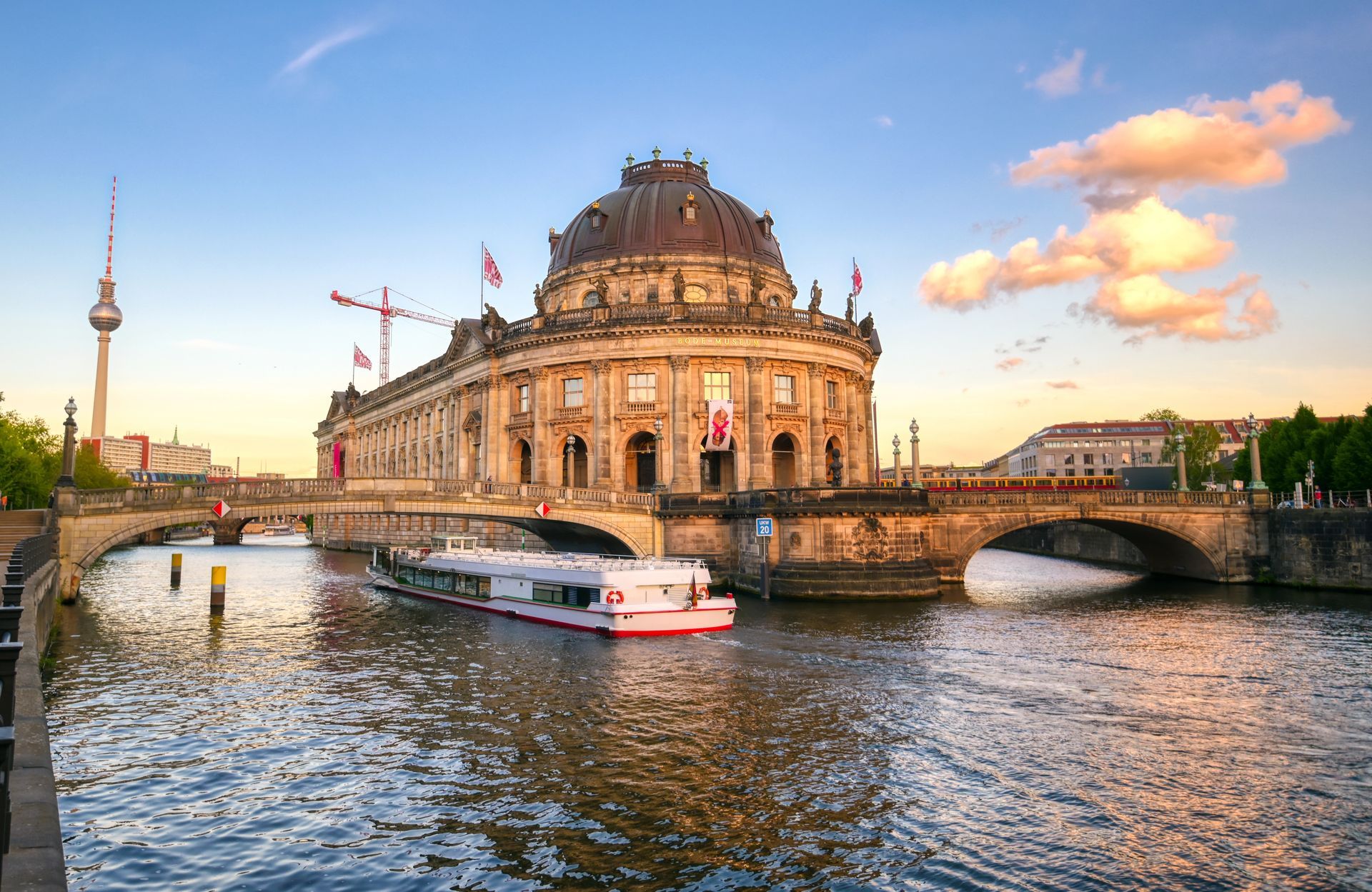 Historic riverside building beside a bridge and boat on a river at sunset, with a city skyline in the background