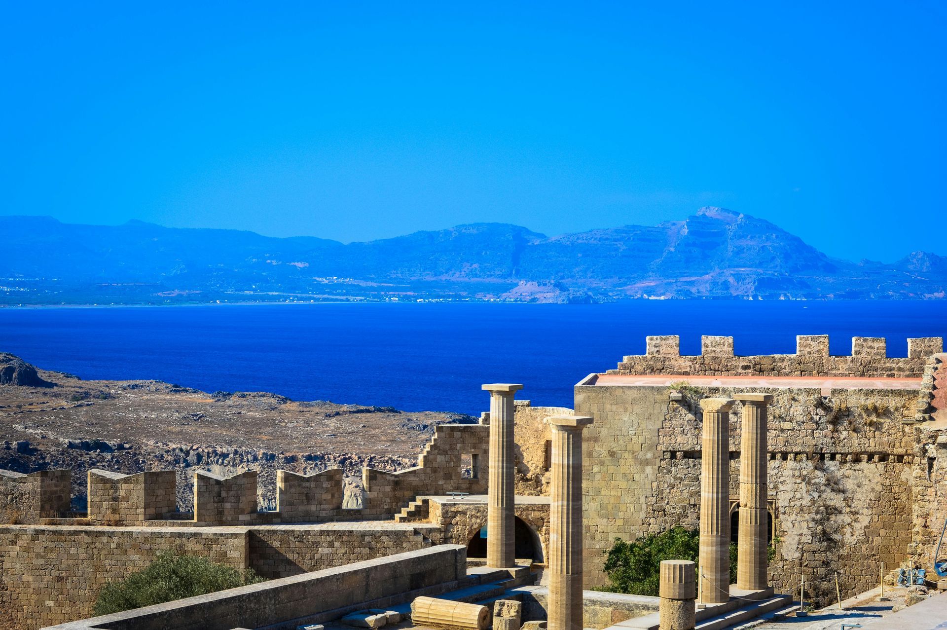 Ancient stone ruins with columns overlooking a bright blue sea and distant mountains.